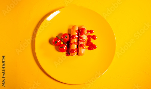 Close up of sausages in sauce with cherry tomatoes on yellow plate. Appetizing frankfurters with small red tomatoes and ketchup on yellow background.