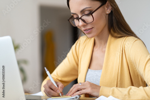 Focused business woman in eyeglasses wear yellow cardigan makes a note in notebook. Office female manager remotely works from home during the second wave of covid-19, coronavirus. Distance job.