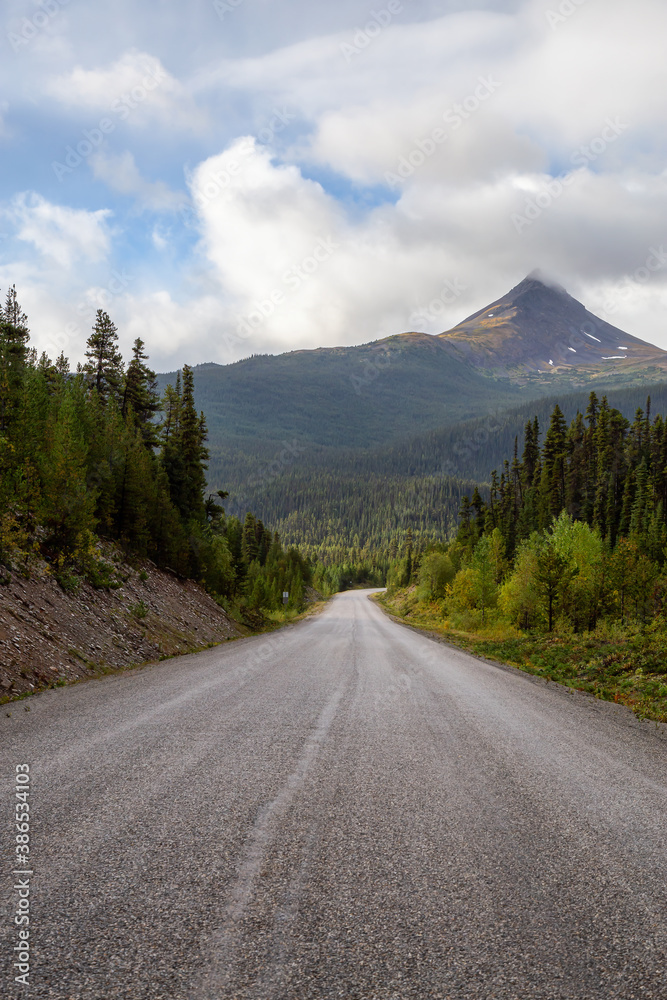 View of Scenic Road surrounded by Trees and Mountains on a Cloudy Fall Morning in Canadian Nature. Taken in Northern British Columbia, Canada. View of Scenic Road surrounded by Trees and Mountains on a Cloudy Fall Morning in Canadian Nature. Taken in Northern British Columbia, Canada.