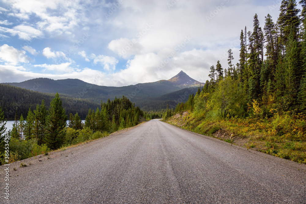 Fototapeta premium View of Scenic Road alongside Lake, surrounded by Trees and Mountains on a Cloudy Fall Morning in Canadian Nature. Taken in Northern British Columbia, Canada.