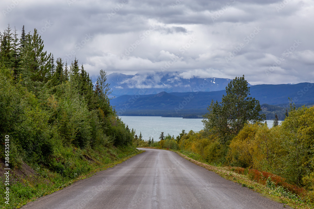 Fototapeta premium View of Scenic Road alongside Lake, surrounded by Trees and Mountains on a Cloudy Fall Day in Canadian Nature. Taken in Northern British Columbia, Canada.