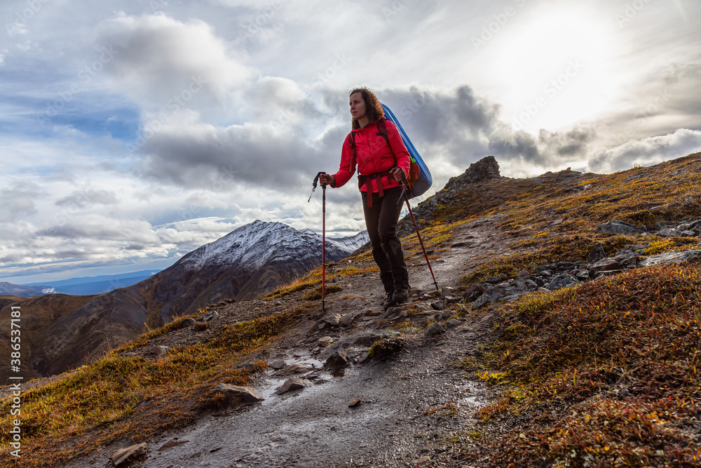 Woman Backpacking along Scenic Hiking Trail surrounded by Mountains in Canadian Nature. Season change from Fall to Winter. Taken in Tombstone Territorial Park, Yukon, Canada. Woman Backpacking along Scenic Hiking Trail surrounded by Mountains in Canadian Nature. Season change from Fall to Winter. Taken in Tombstone Territorial Park, Yukon, Canada.