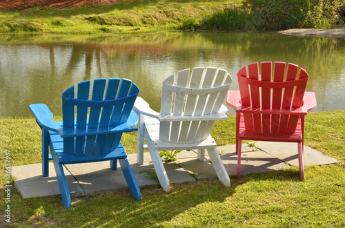 Tranquil scene with patriotic fourth of july theme. Three colorful red, white, and blue lawn chairs overlooking small pond.