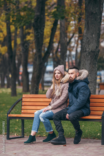 A guy and a girl are resting on a bench in an autumn park. A loving couple in jackets sits on a bench in the main park © Дмитрий Скорина