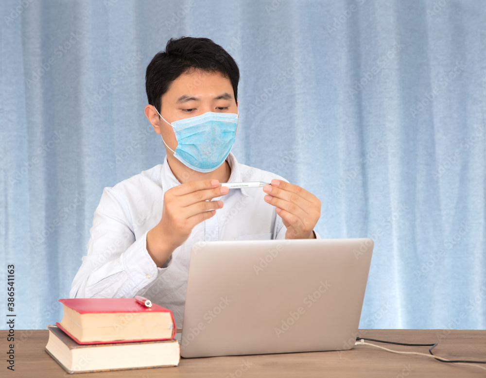 Man wearing a mask at his desk looking at a thermometer Man wearing a mask at his desk looking at a thermometer