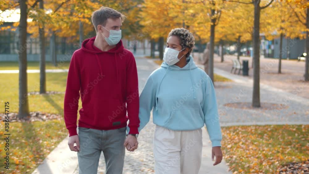Young diverse couple walking hand in hand wearing medical mask in autumn park