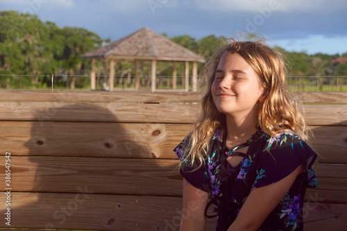 Girl smiling with eyes closed on a wooden bench, feeling the sun and enjoying the nature view