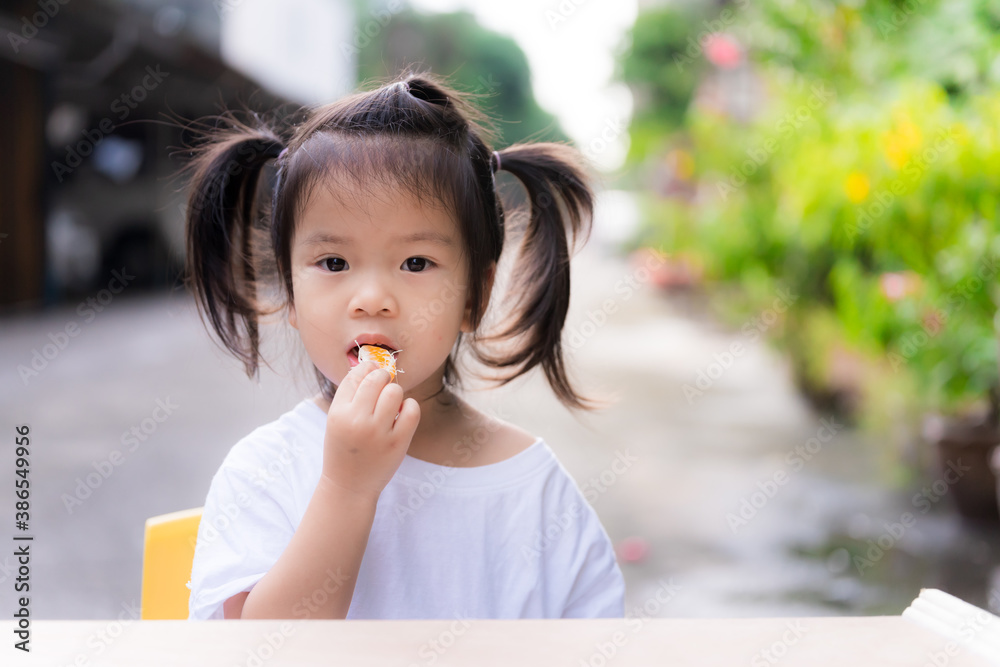 Adorable girl is eating oranges. It is difficult for children to eat fruit. Child wear white shirts. Little wooden table in front. Cute kid tied up with two black hair. Toddler are 3 years old. Adorable girl is eating oranges. It is difficult for children to eat fruit. Child wear white shirts. Little wooden table in front. Cute kid tied up with two black hair. Toddler are 3 years old.
