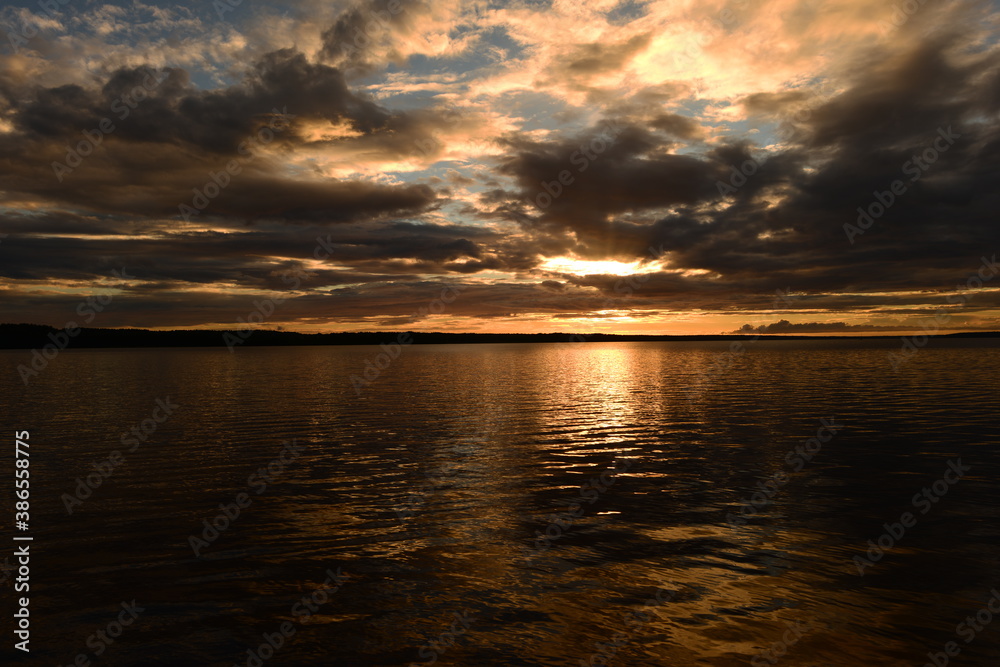 Fototapeta premium Glowing sunlight on the cloudy twilight sky is reflected by colorful light and shadows on the lake water