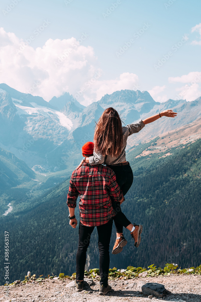 Naklejka premium Beautiful couple among the mountains. A couple in love in the mountains, back view. The man lifted the woman into his arms. The couple travels among the mountains. Hiking in the mountains. Copy space