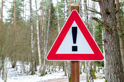 Winter road in forest on background. Road sign triangular shape with exclamation mark. Red with white attention traffic sign.