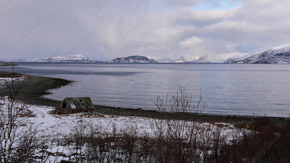 Collapsed old wooden shed at the shore of Altafjord, Troms og Finnmark, Norway with bare trees and snow-covered mountains on cloudy day in winter time.