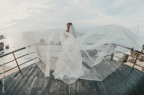 Young woman in long white flying dress and veil standing over the ocean beach in Maldives. Bride rear view, gown fabric fly on wind. Tropical wedding on Bali.