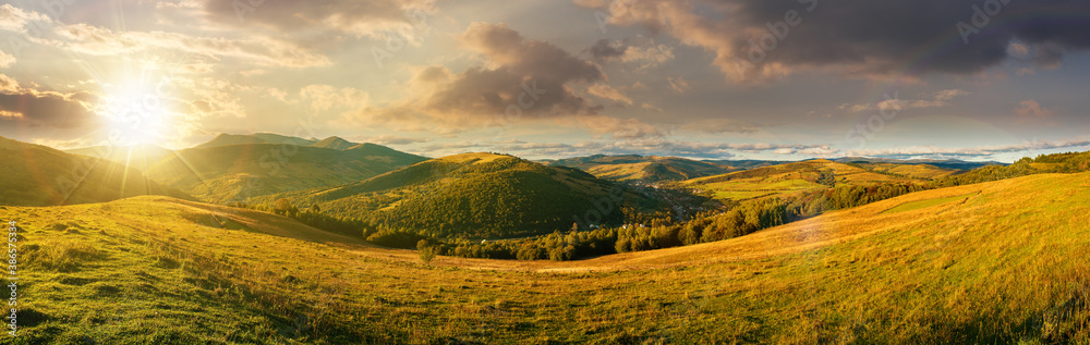 mountainous countryside landscape at sunset. panorama of a grassy rural field on the hill in evening light. village in the distant valley. clouds on the sky