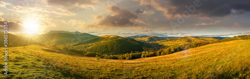 mountainous countryside landscape at sunset. panorama of a grassy rural field on the hill in evening light. village in the distant valley. clouds on the sky
