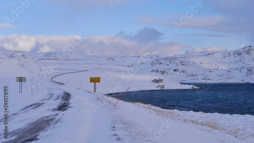 Icy road E69 with road sign displaying distance to Nordkapp (45km) near Honningsvåg, Norway, Scandinavia with red snowplow markings, remote house, snow-covered landscape and arctic sea in winter time.