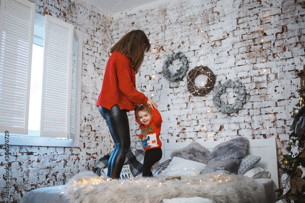 Mom with daughter in red warm sweaters are jumping on the bed. Happy motherhood. Warm family relationships. Christmas and New Year's interior. Love. Family concept. Mom with daughter in red warm sweaters are jumping on the bed. Happy motherhood. Warm family relationships. Christmas and New Year's interior. Love. Family concept.