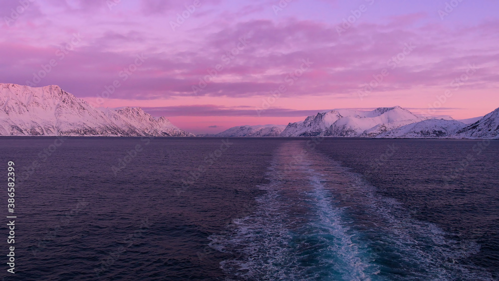 Stunning panorama view from the stern of a cruise ship with the vessel's wake in the arctic sea, snow-covered mountains and a beautiful purple colored sky after sunset in winter time. - obrazy, fototapety, plakaty 
