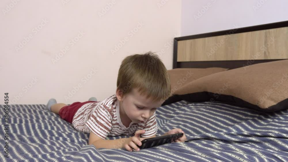 Little boy plays in a smartphone while lying on a big bed.4k Little boy plays in a smartphone while lying on a big bed.4k