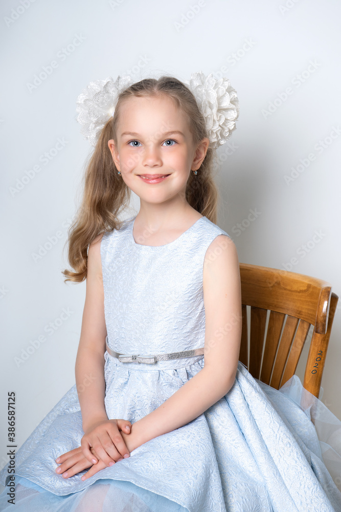 Portrait of an elegant seven-year-old girl in a blue festive dress with hairstyles with bows, sits on a chair on a light background. Retro style Portrait of an elegant seven-year-old girl in a blue festive dress with hairstyles with bows, sits on a chair on a light background. Retro style