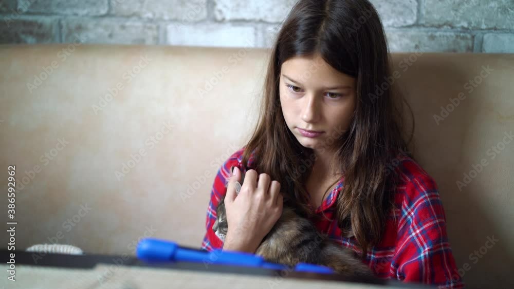 Girl wearing checkered pajamas sitting on sofa and stroking cat while watching tablet. Child entertaining herself with gadget. Concept of leisure Girl wearing checkered pajamas sitting on sofa and stroking cat while watching tablet. Child entertaining herself with gadget. Concept of leisure