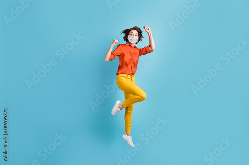 Full length photo of crazy lady jump fists rejoicing wear mask orange shirt yellow trousers sneakers isolated blue color background