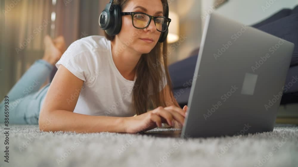 Woman is lying on the floor, working on a laptop and listens to music on headphones. Concept of remote work. Woman is lying on the floor, working on a laptop and listens to music on headphones. Concept of remote work.
