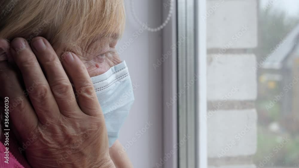 Old Woman In Protective Mask Looks Out Window And Holds Her Sick Head In Hands Old Woman In Protective Mask Looks Out Window And Holds Her Sick Head In Hands