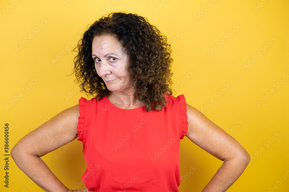 Middle age woman wearing casual shirt standing over isolated yellow background skeptic and nervous, disapproving expression on face with arms in waist Middle age woman wearing casual shirt standing over isolated yellow background skeptic and nervous, disapproving expression on face with arms in waist