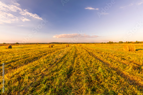 Scenic view at picturesque burning sunset in a green shiny field with hay stacks, bright cloudy sky , trees and golden sun rays, summer valley landscape © Yaroslav