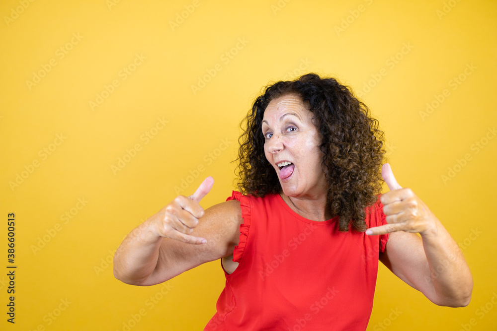 Middle age woman wearing casual shirt standing over isolated yellow background shouting with crazy expression doing rock symbol with hands up Middle age woman wearing casual shirt standing over isolated yellow background shouting with crazy expression doing rock symbol with hands up
