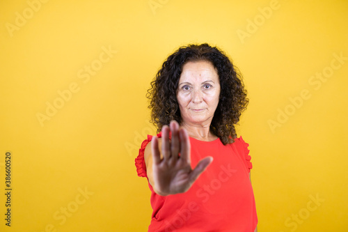 Middle age woman wearing casual shirt standing over isolated yellow background serious and doing stop sing with palm of the hand.