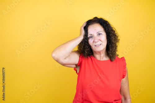 Middle age woman wearing casual shirt standing over isolated yellow background putting one hand on her head smiling like she had forgotten something