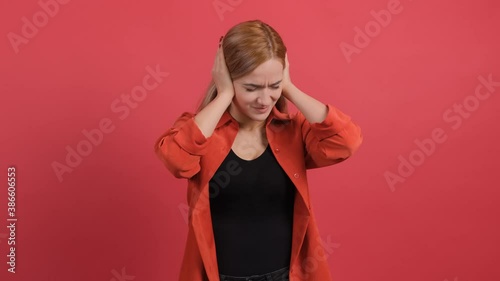 Close up of a confused woman holding her hands to her ears and nodding, isolated over red background. Portrait of young serious girl.