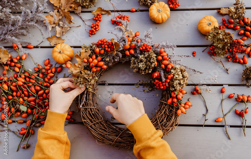 Girl making floral autumn door wreath using colorful rosehip berries, rowan, dry flowers and pumpkins. Fall flower decoration workshop, florist at work.