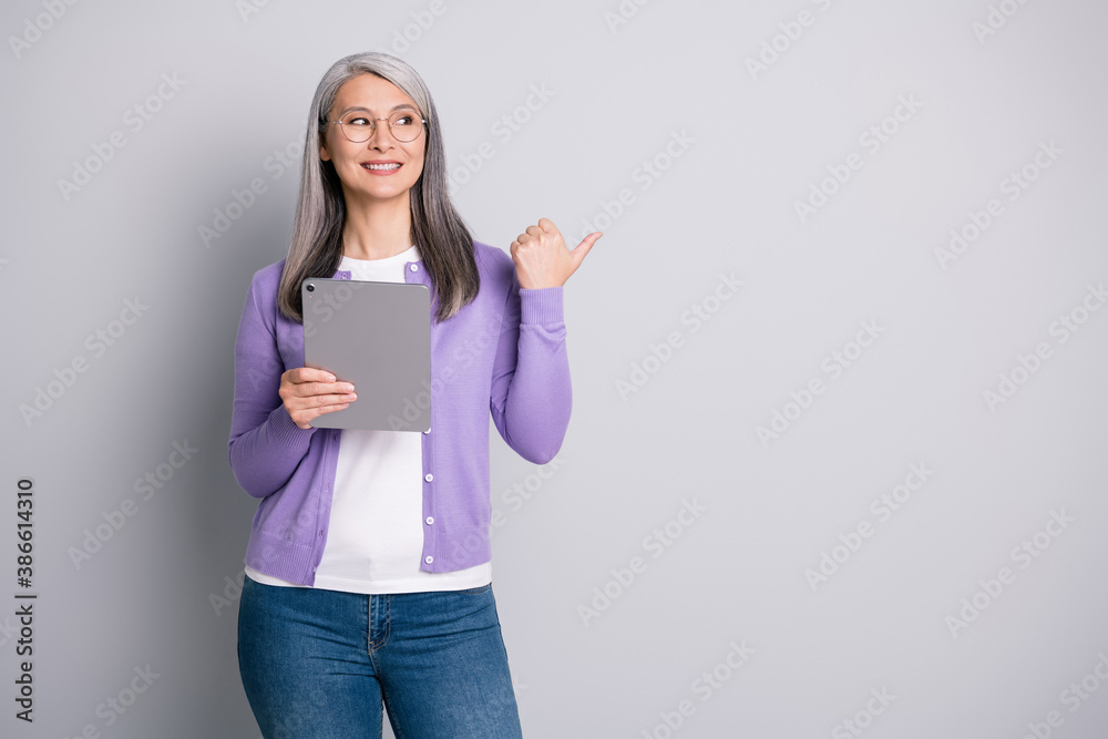 Photo portrait of senior lady using laptop for reading information pointing showing with thumb looking at side wearing purple cardigan isolated on grey color background Photo portrait of senior lady using laptop for reading information pointing showing with thumb looking at side wearing purple cardigan isolated on grey color background