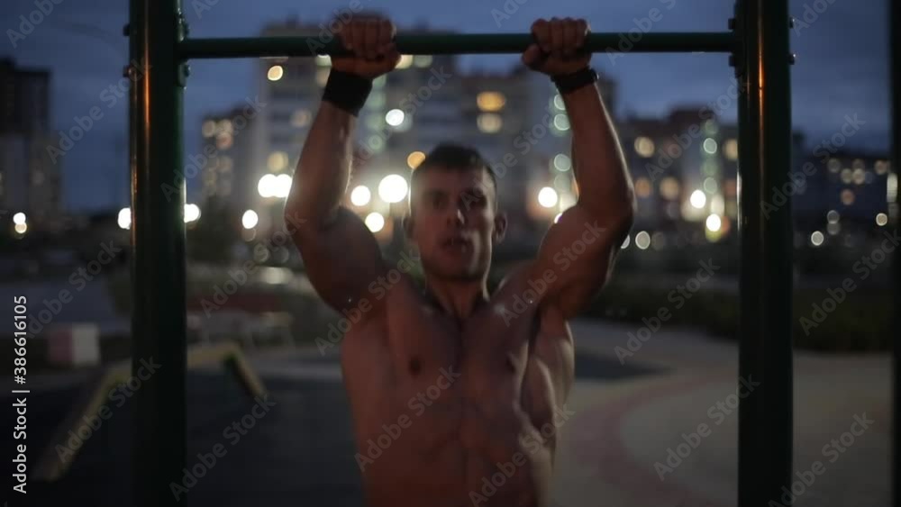 A young bodybuilder without a shirt works out on a city sports field on a summer evening and pulls up on a horizontal bar. Front view. Close-up. Blurred background A young bodybuilder without a shirt works out on a city sports field on a summer evening and pulls up on a horizontal bar. Front view. Close-up. Blurred background