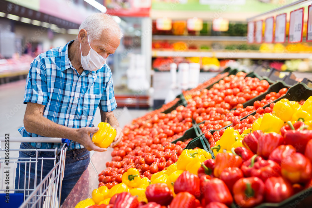 elderly caucasian man in mask with covid protection choosing tomatoes in vegetable section of supermarket
