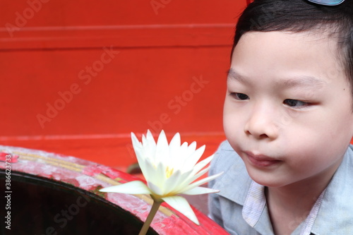 an asian Chinese cute young boy in grey shirt is looking at and smell the scent of  the white lotus flower in big water jar with red cement wall background photo