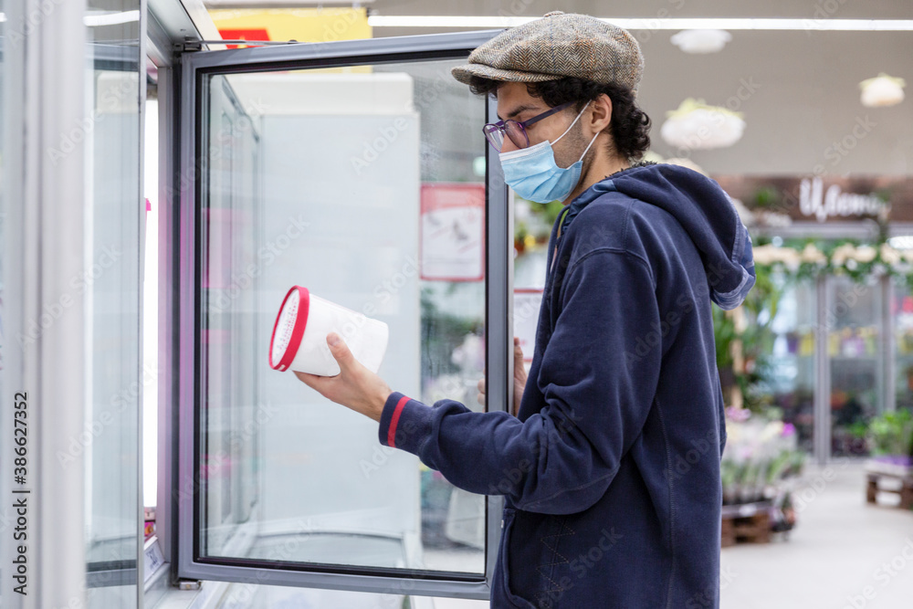 A young man in a medical mask chooses frozen food in a supermarket. Coronavirus pandemic. A young man in a medical mask chooses frozen food in a supermarket. Coronavirus pandemic.