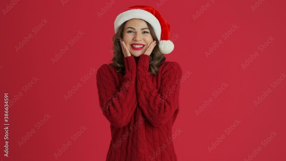 A surprising woman wearing a warm red sweater and christmas hat is looking to the camera standing isolated over red background
