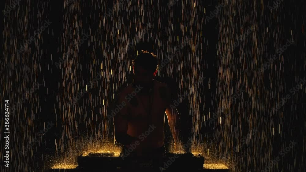 Silhouette of a young man mixing music behind a DJ set in a dark studio in the pouring rain. Water droplets drum on the surface with the mixer controller in slow motion. Close up.