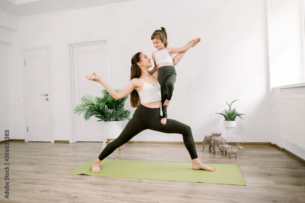Attractive young Caucasian woman and her cute little daughter are smiling while doing yoga together at home. Pretty mom stands in Warrior One position and holds her little child girl Attractive young Caucasian woman and her cute little daughter are smiling while doing yoga together at home. Pretty mom stands in Warrior One position and holds her little child girl