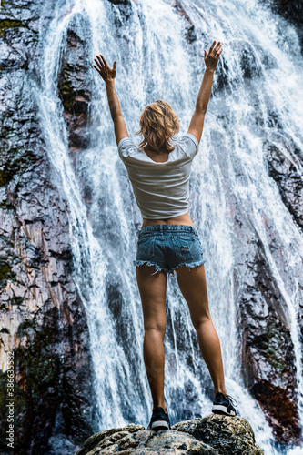 Young caucasian slender woman tourist in casual clothes and sneakers stands on a rock and look at waterfall 