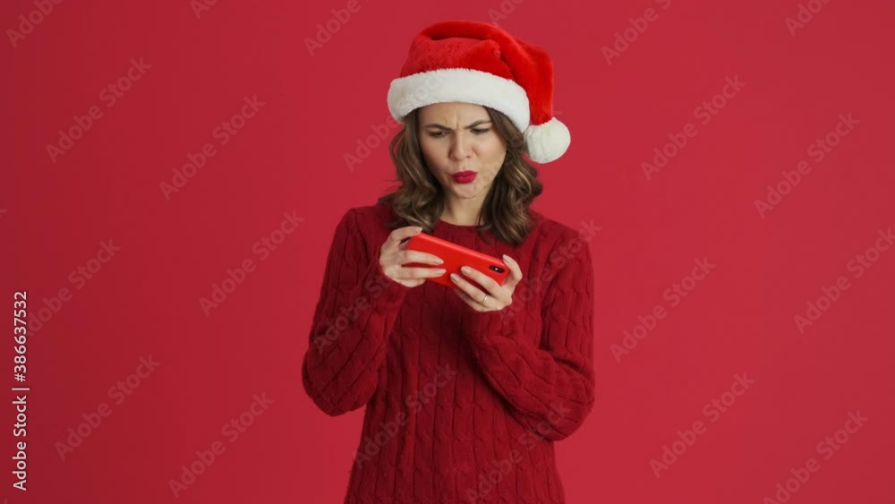 A smiling woman wearing a warm red sweater and christmas hat is playing games on her smartphone standing isolated over red background A smiling woman wearing a warm red sweater and christmas hat is playing games on her smartphone standing isolated over red background