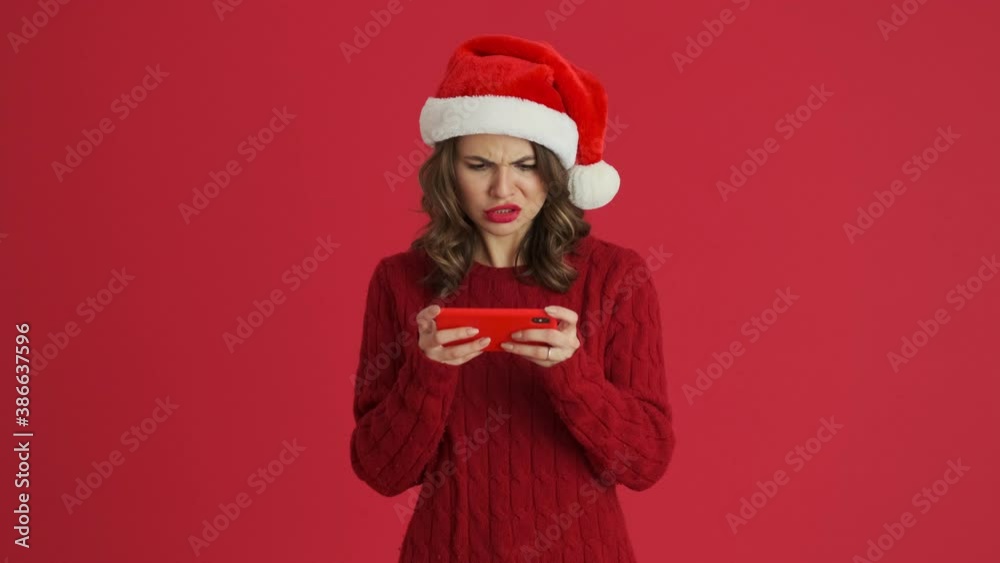 An emotional woman wearing a warm red sweater and christmas hat is losing the game on her smartphone standing isolated over red background An emotional woman wearing a warm red sweater and christmas hat is losing the game on her smartphone standing isolated over red background