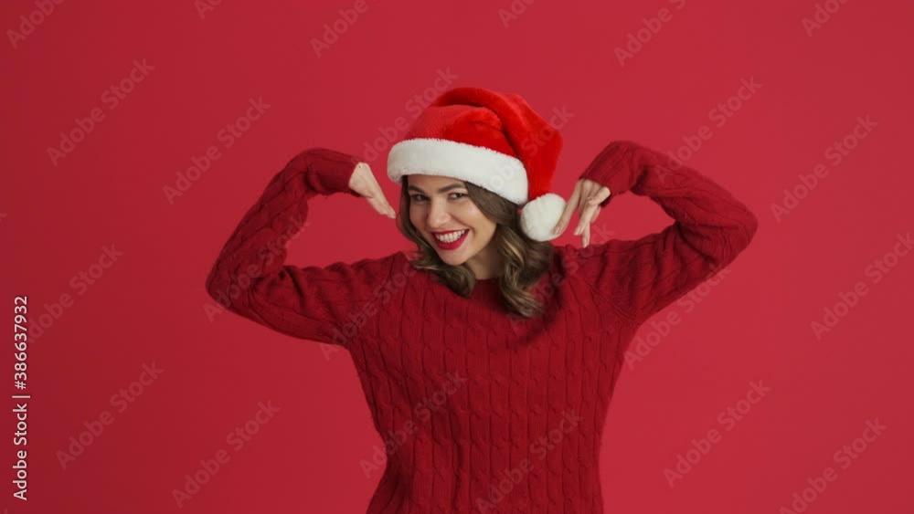 A happy woman wearing a warm red sweater and christmas hat is dancing isolated over red background