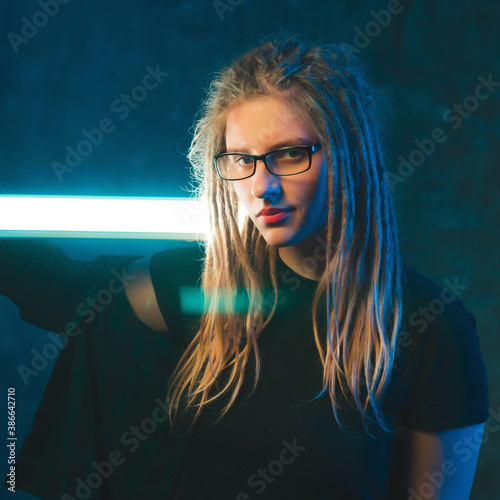 Girl with dreadlocks in blue neon lighting on dark background. Stylish young woman with strip lamp in futuristic environment