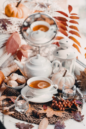 A cup of hot tea with lemon in an early autumn morning by the window. A stack of sweaters, yellow leaves, pumpkin, candles. Decor. Autumn concept of warm drink on a cold day.