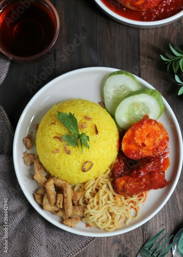 Top view of Nasi Kuning, Indonesian popular traditional  yellow rice served with chicken and eggs balado, noodle,  tempeh, and cucumber slices. Dark wooden background photo